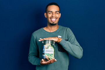 Young african american man holding charity jar with south africa rand banknotes sticking tongue out happy with funny expression.