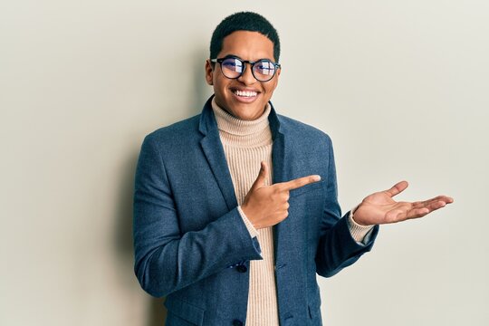 Young handsome hispanic man wearing elegant clothes and glasses amazed and smiling to the camera while presenting with hand and pointing with finger.