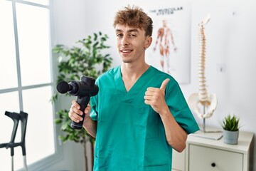 Fototapeta na wymiar Young caucasian physio man holding muscle percusion gun at the clinic smiling happy and positive, thumb up doing excellent and approval sign
