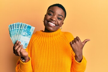 Young african american woman holding 100 brazilian real banknotes pointing thumb up to the side...