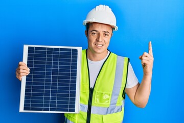 Handsome young man holding photovoltaic solar panel surprised with an idea or question pointing finger with happy face, number one