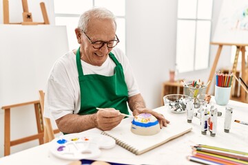 Senior grey-haired artist man smiling happy painting pottery sitting on the table at art studio.