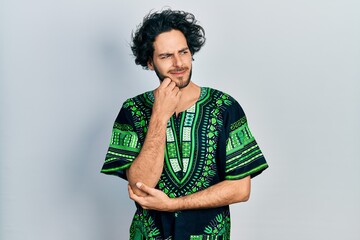 Handsome hispanic man wearing traditional african clothes serious face thinking about question with hand on chin, thoughtful about confusing idea