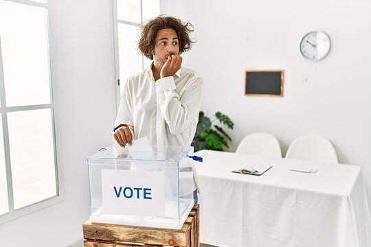 Young hispanic man voting putting envelop in ballot box looking stressed and nervous with hands on mouth biting nails. anxiety problem.