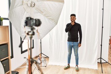 Young hispanic man with beard posing as model at photography studio looking confident at the camera with smile with crossed arms and hand raised on chin. thinking positive.