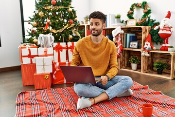 Arab young man using laptop sitting by christmas tree smiling looking to the side and staring away thinking.