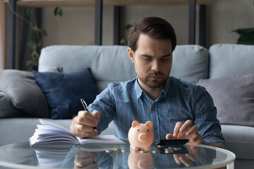 Serious millennial man calculating savings, counting budget after paying bill, taxes, mortgage fees, making notes at table with piggy bank, cash. Homeowner doing bookkeeping work. Finance concept