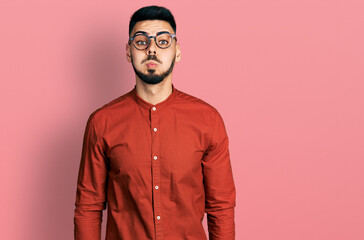 Young hispanic man with beard wearing business shirt and glasses puffing cheeks with funny face. mouth inflated with air, crazy expression.