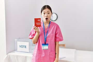 Young asian nurse woman at political campaign election holding swiss passport scared and amazed with open mouth for surprise, disbelief face