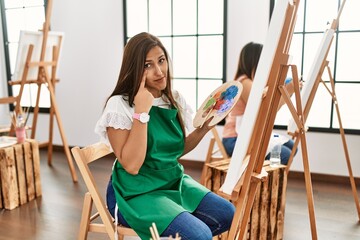 Young hispanic artist women painting on canvas at art studio pointing to the eye watching you gesture, suspicious expression