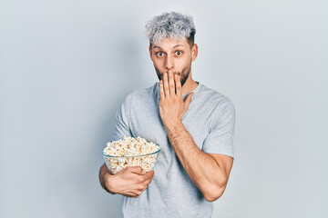 Young hispanic man with modern dyed hair eating popcorn covering mouth with hand, shocked and...