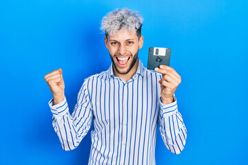 Young hispanic man with modern dyed hair holding floppy disk screaming proud, celebrating victory...
