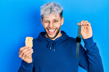 Young hispanic man with modern dyed hair holding wireless earphones and smart watch winking looking...