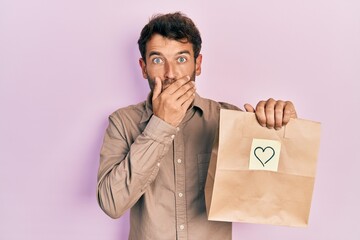 Handsome man with beard holding delivery paper bag with heart reminder shocked covering mouth with...