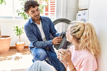 Young couple smiling happy drinking coffee while doing laundry at home.