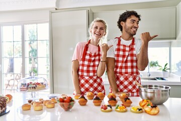 Couple of wife and husband cooking pastries at the kitchen smiling with happy face looking and pointing to the side with thumb up.