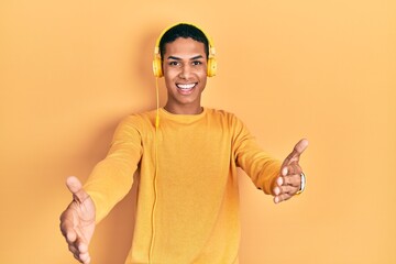 Young african american guy listening to music using headphones smiling cheerful offering hands giving assistance and acceptance.