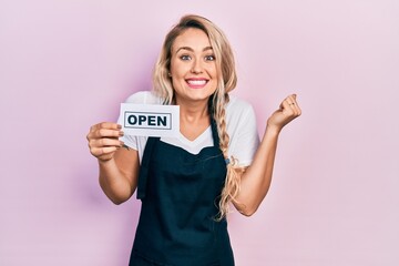 Beautiful young blonde woman wearing waitress apron holding open banner screaming proud, celebrating victory and success very excited with raised arm