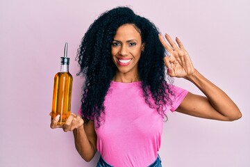 Middle age african american woman holding olive oil can doing ok sign with fingers, smiling...