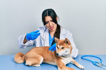 Beautiful hispanic veterinarian woman putting vaccine to puppy dog in shock face, looking skeptical and sarcastic, surprised with open mouth
