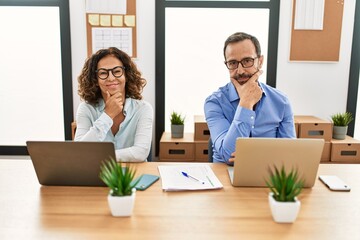 Fototapeta premium Middle age hispanic woman and man sitting with laptop at the office looking confident at the camera with smile with crossed arms and hand raised on chin. thinking positive.