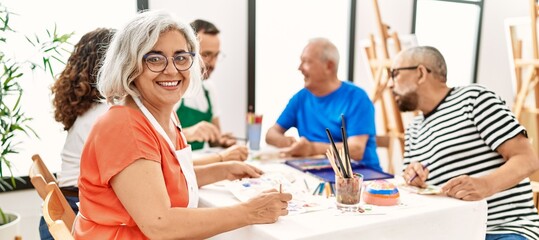 Group of middle age draw students sitting on the table drawing at art studio. Woman smiling happy looking to the camera. - obrazy, fototapety, plakaty