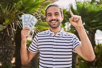 Young hispanic man holding dollars close to face smiling with an idea or question pointing finger...