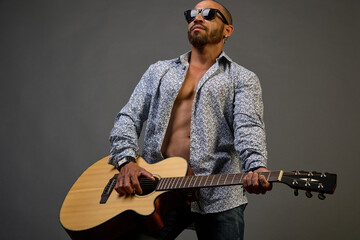 Cool muscular Latin American in sunglasses and a shirt plays music on an acoustic guitar on a gray...