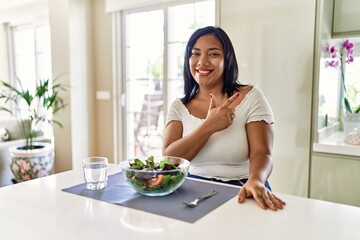 Obraz na płótnie Canvas Young hispanic woman eating healthy salad at home cheerful with a smile of face pointing with hand and finger up to the side with happy and natural expression on face