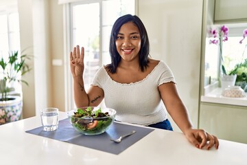 Obraz na płótnie Canvas Young hispanic woman eating healthy salad at home showing and pointing up with fingers number four while smiling confident and happy.