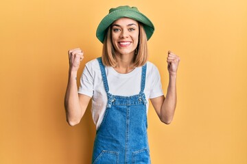 Young caucasian blonde woman wearing denim jumpsuit and hat with 90s style celebrating surprised...