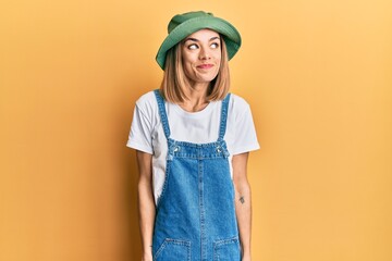 Young caucasian blonde woman wearing denim jumpsuit and hat with 90s style smiling looking to the...