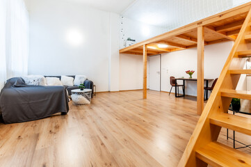 Bright loft apartment with large window, wooden floor and built-in wooden staircase. The room has decorative coffee table, big corner sofa with pillows, wall mounted lights and set dining table.