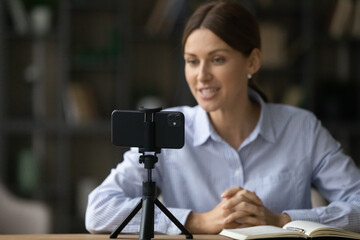 Focus on smartphone on tripod, smiling young woman recording video, sitting at desk, popular blogger influencer shooting content for social network, business coach mentor leading online lesson