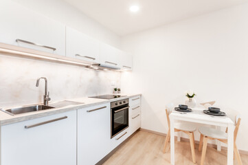 Kitchen at home with marble on wall,white cabinets,regular appliances and stonelike work top. Kitchen is decorated with tiny plants and illuminated with light strip. On right is set a dinning table.