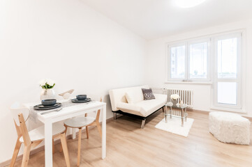 Spacious living room with sofa decorated in modern minimalist style. Wooden materials and white objects predominate in room. At back is a large balcony window. In the foreground a small dining table.