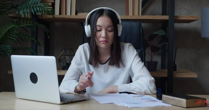 Focused young asian student wearing wireless headphones, studying on online courses, using paper notes. Motivated millennial internet teacher giving language class by conference video call.