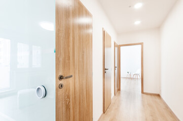 Fototapeta na wymiar Hallway with contemporary wooden room door designs, wooden floor and opened entrance in the end. All doors have a wooden texture and fittings made of brown matt stainless steel. 