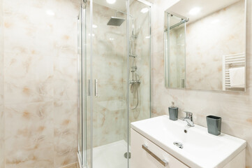 Contemporary bathroom with shower and sink with white cabinet under it and mirror on wall. All walls are lined with marble in beige shades. Whole space gives modern and warm impression.