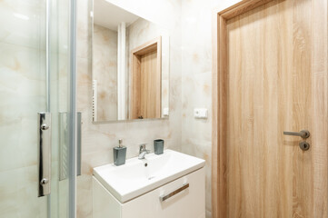 Fototapeta na wymiar Contemporary bathroom with glassy walled shower and sink with white cabinet under it and mirror on wall. All walls are lined with marble in beige shades. Door are made of wood.