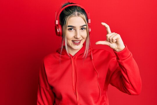 Young modern girl wearing gym clothes and using headphones smiling and confident gesturing with hand doing small size sign with fingers looking and the camera. measure concept.