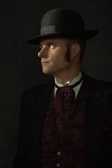 Fototapeta na wymiar Shadowy portrait of a young man in stylish vintage Victorian attire in front of a dark gray wall.