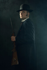 Shadowy portrait of a young man in vintage Victorian attire holding a rifle in a smoky room in...