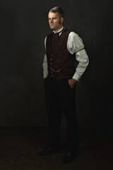 Shadowy portrait of a young man in vintage Victorian attire in a room in front of a dark gray wall.