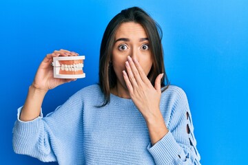 Young hispanic girl holding denture covering mouth with hand, shocked and afraid for mistake....