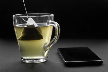Green tea in a pyramid tea bag is brewed in hot water in a transparent cup next to a smartphone. The concept of relaxation and tea drinking together with a mobile gadget