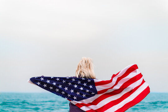 Caucasian blonde woman holding american flag leaning against back and looking at sea. Usa flag for independence day