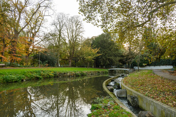Outdoor scenery of walking way along Jeker canal and historical city wall at Monseigneur Nolenspark, city public park, in Autumn season, in Maastricht, Netherlands during evening sunset time.
