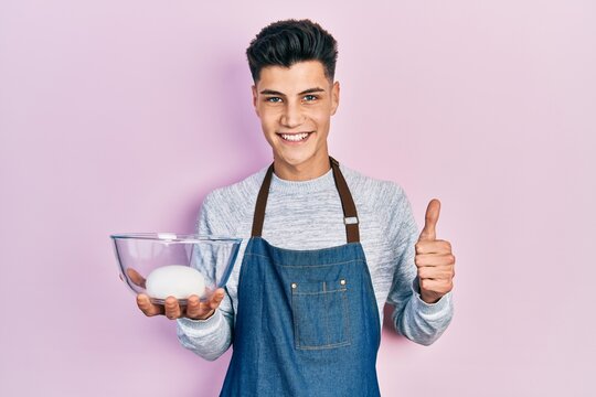 Young hispanic man wearing apron holding bowl of bread dough smiling happy and positive, thumb up doing excellent and approval sign