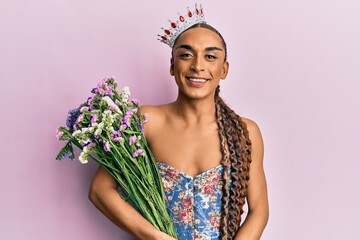 Hispanic man wearing make up and long hair wearing princess crown holding bouquet of flowers looking positive and happy standing and smiling with a confident smile showing teeth © Krakenimages.com